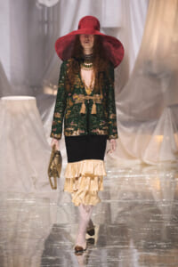 Model walks the runway in a green sequined jacket, layered beige ruffled skirt, and a wide red hat.