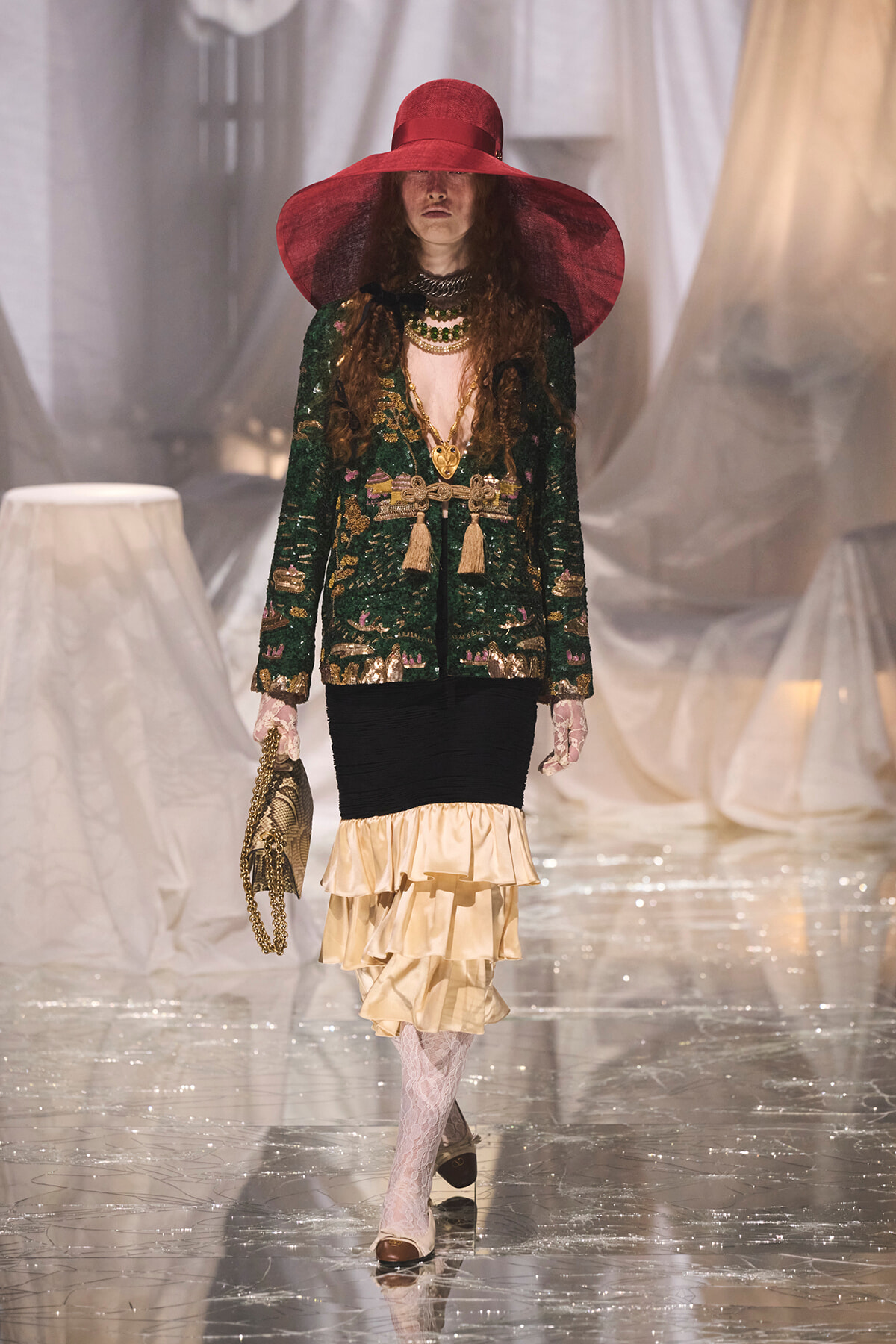 Model walks the runway in a green sequined jacket, layered beige ruffled skirt, and a wide red hat.
