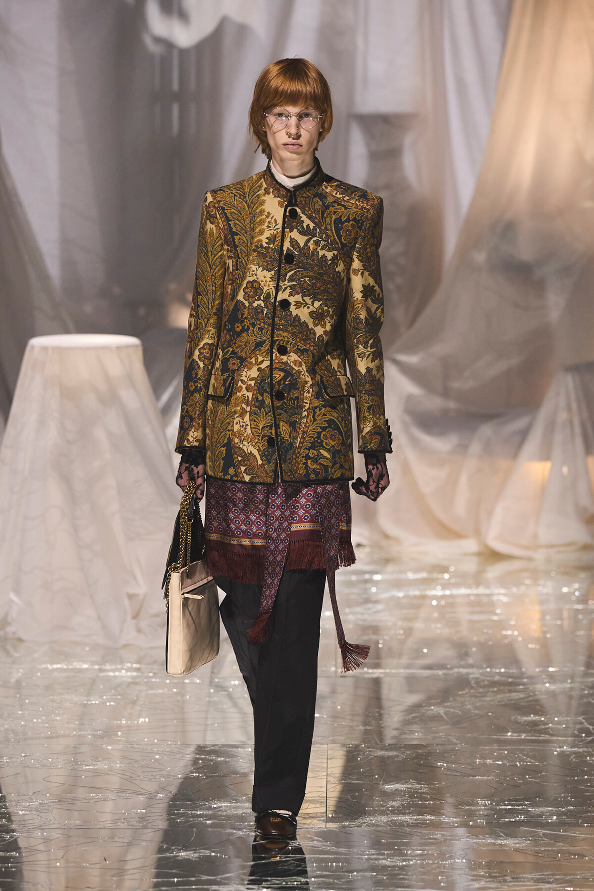 Model on a glossy runway wearing a gold-brown ornate floral blazer, black trousers, and round glasses, carrying a beige handbag.