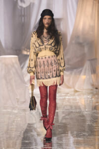 Model on a runway wearing a cream beaded blouse with black embroidery, a pale pink fringe skirt, red lace tights, a black veil cap, and a small black handbag with a gold chain.