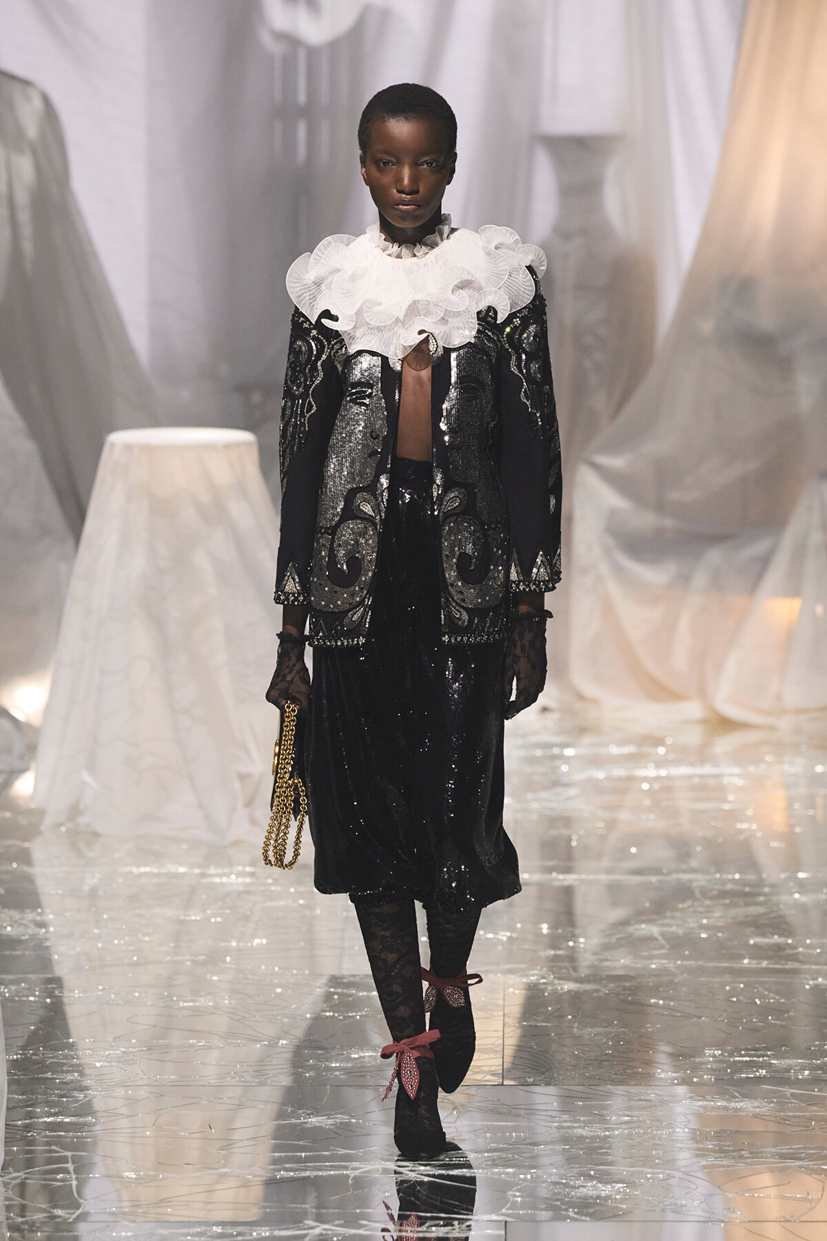 Model walks a fashion runway in a black sequined coat-dress with a large white ruffled collar, lace gloves, and a gold chain purse. The glossy floor and pale drapes form the backdrop.