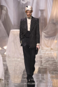 Male model on runway wearing a black polka-dot blazer, white shirt, bow tie, and black trousers; accessorized with a white turban and lace glove, holding a clutch.