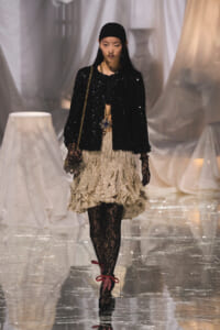 Model walking a runway in a black sequined jacket, beige polka-dot ruffled skirt, and black lace tights, carrying a gold chain bag.