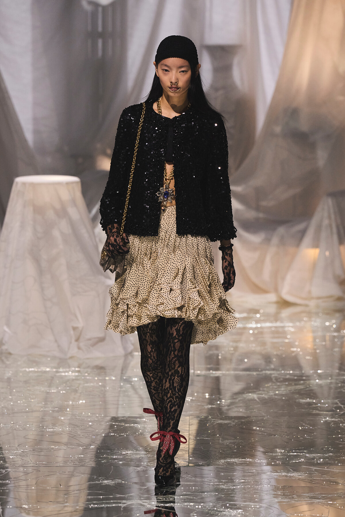 Model walking a runway in a black sequined jacket, beige polka-dot ruffled skirt, and black lace tights, carrying a gold chain bag.