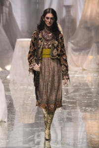 Model walking runway in a heavily embroidered velvet jacket, layered jewelry, and a patterned dress with mustard belt and snakeskin boots.
