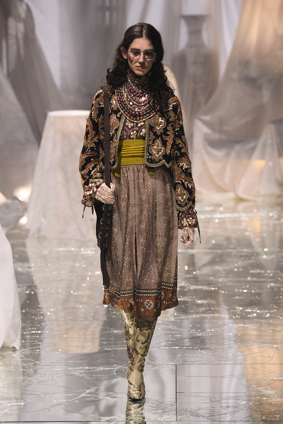 Model walking runway in a heavily embroidered velvet jacket, layered jewelry, and a patterned dress with mustard belt and snakeskin boots.