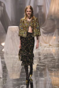 Model walking a runway in a gold embroidered jacket over a black floral, tiered skirt with lace tights and gold shoes.