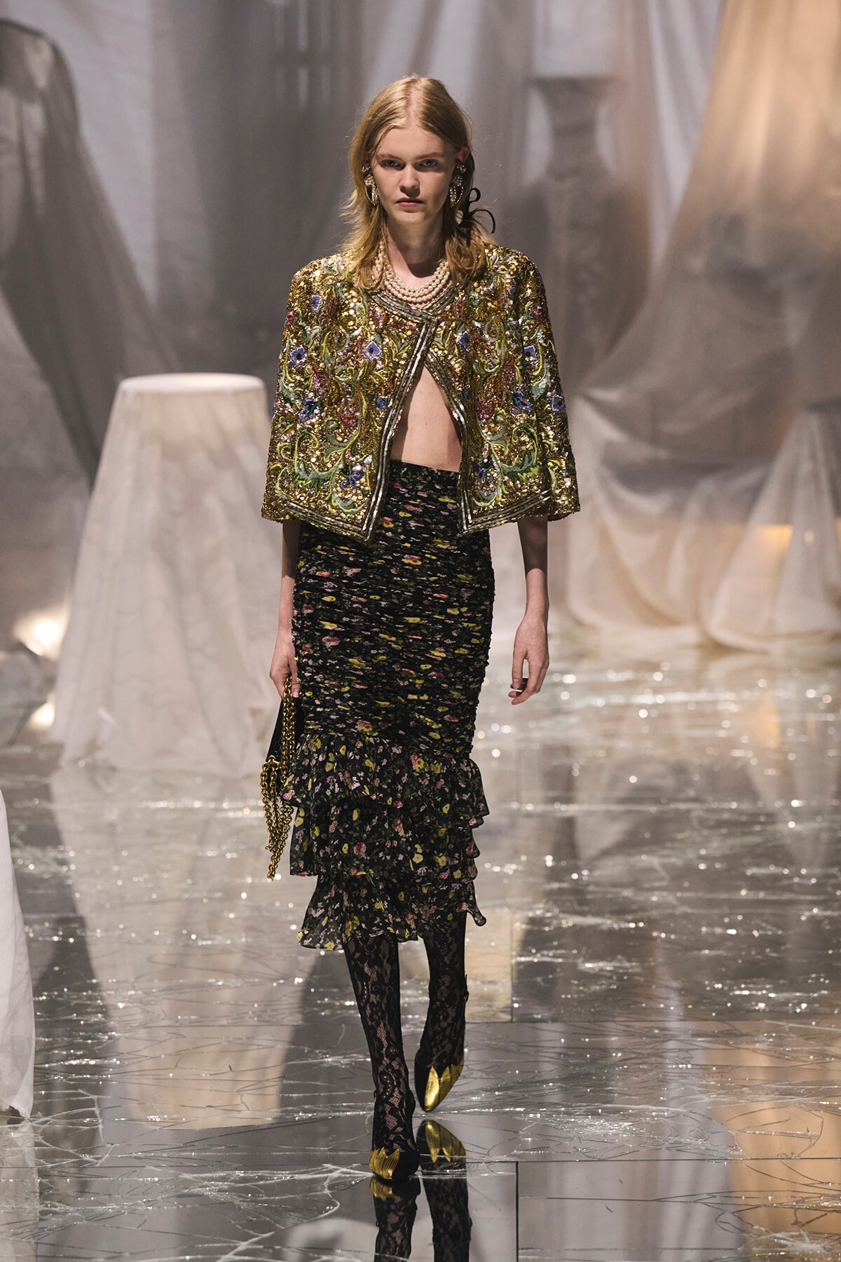 Model walking a runway in a gold embroidered jacket over a black floral, tiered skirt with lace tights and gold shoes.