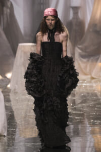 Model on a runway wearing a strapless black polka-dot gown with tiered ruffles, veil, and a pink satin headscarf.