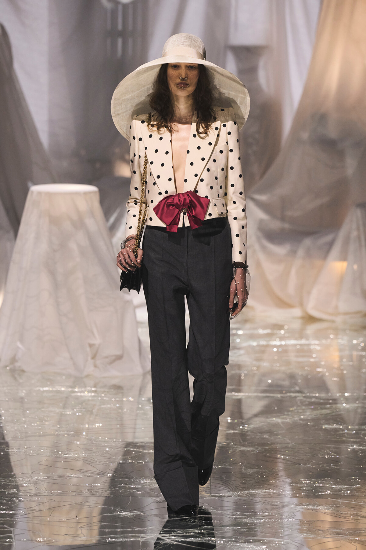 Model walks the runway in a white polka-dot blazer with a pink bow at the waist, a wide-brim hat, and dark trousers, carrying a chain-strap bag.