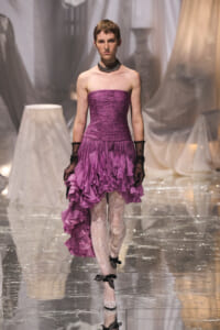 Runway model in a strapless purple ruffled dress with long black lace gloves on a metallic catwalk.