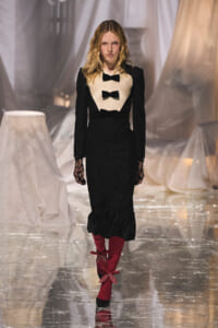 Model walking the runway in a black long-sleeve dress with beige bib and three black bows, plus lace gloves and red knee-high boots with bows.