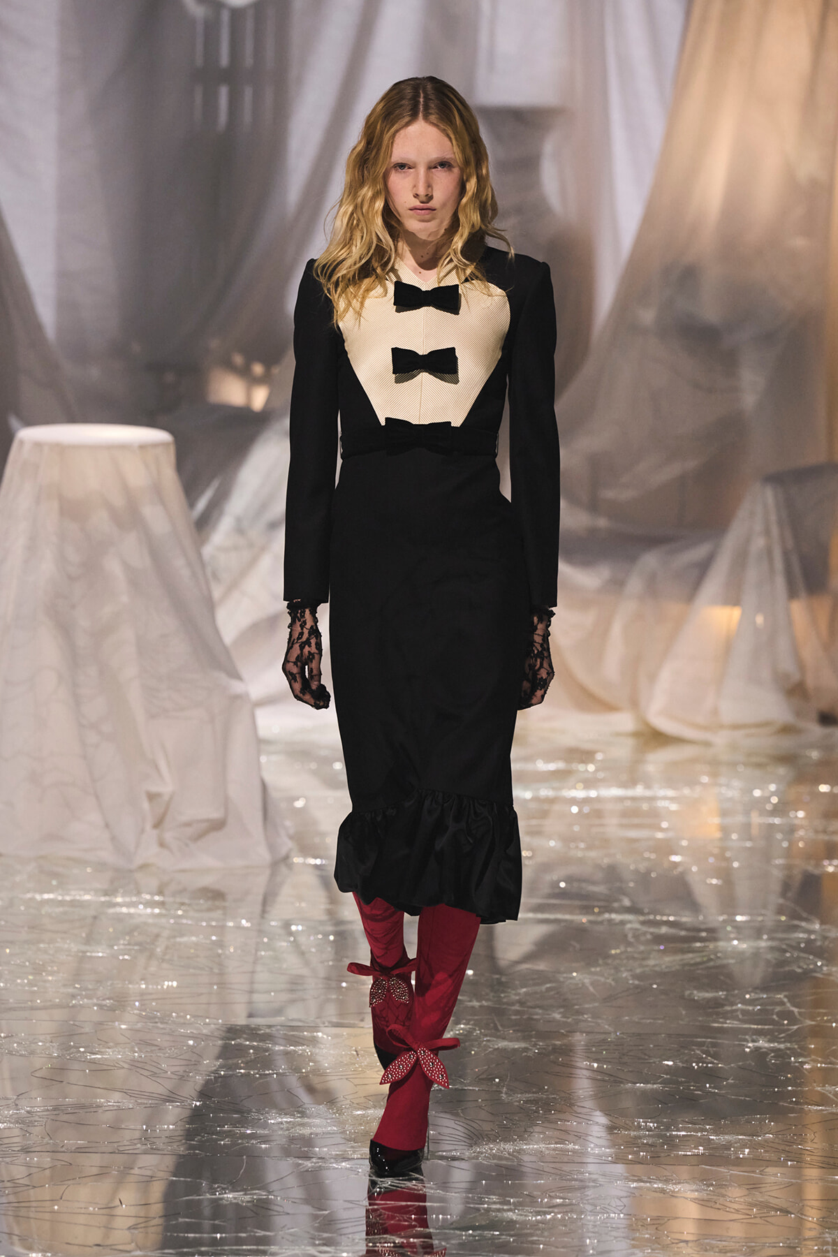 Model walking the runway in a black long-sleeve dress with beige bib and three black bows, plus lace gloves and red knee-high boots with bows.