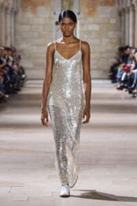 Model walking a runway in a silver sequin slip dress with thin straps at a fashion show, surrounded by an audience.
