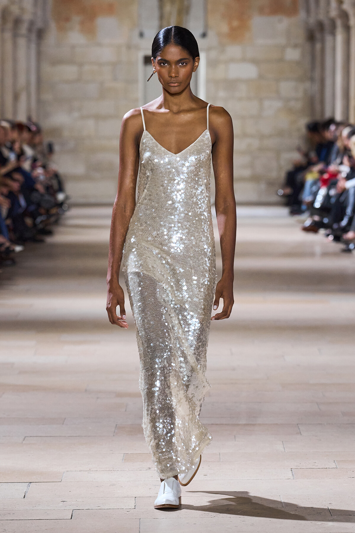 Model walking a runway in a silver sequin slip dress with thin straps at a fashion show, surrounded by an audience.