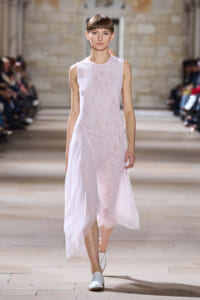Model walking a runway in a pale pink sleeveless dress with a flowing skirt; audience blurred in background.”] ,