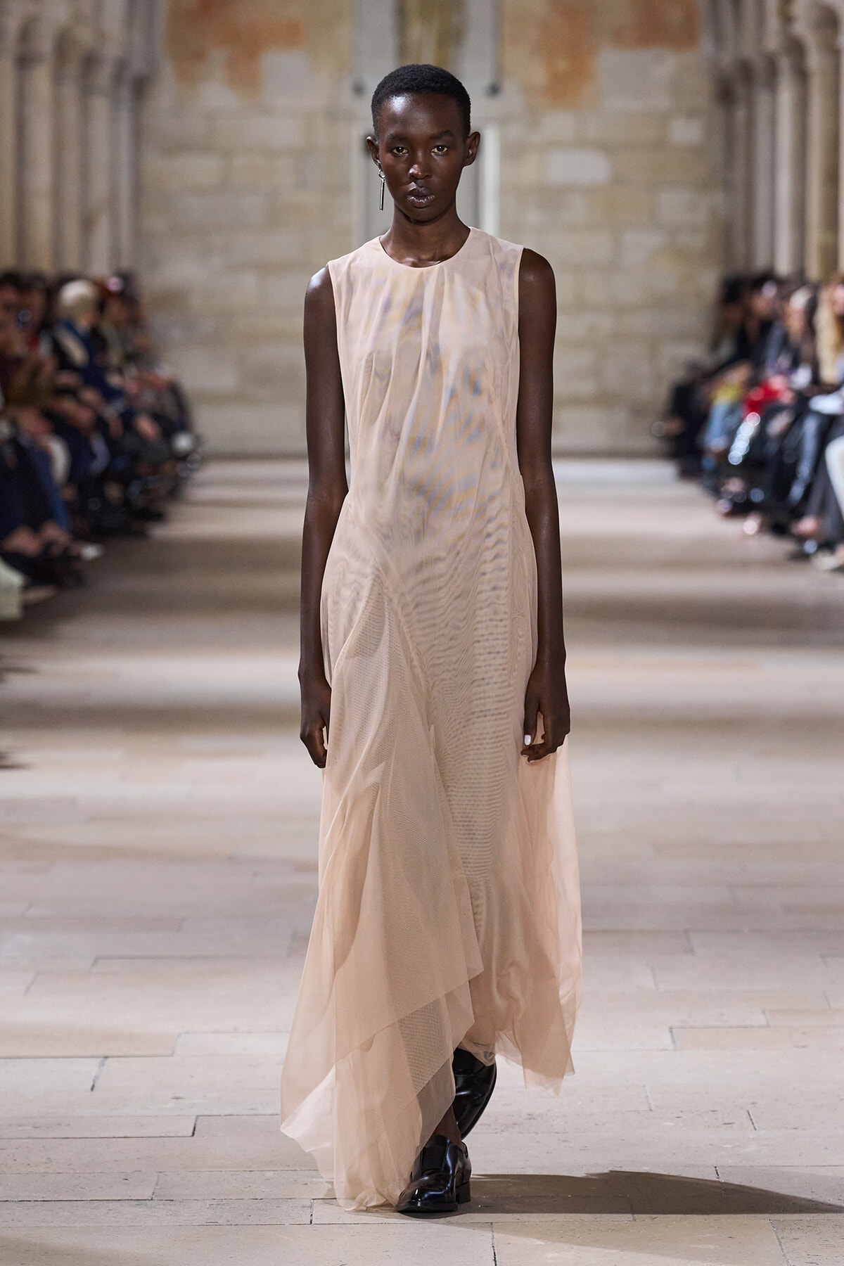 Model walking a peach sheer sleeveless gown on a runway during a fashion show, with seated audience on both sides.
