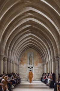 Model walks down a stone Gothic arcade runway in an orange-brown outfit, audience on both sides.
