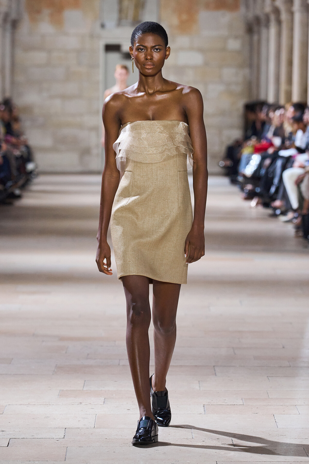 Model wearing a beige strapless mini dress with sheer ruffle layers, walking a runway.