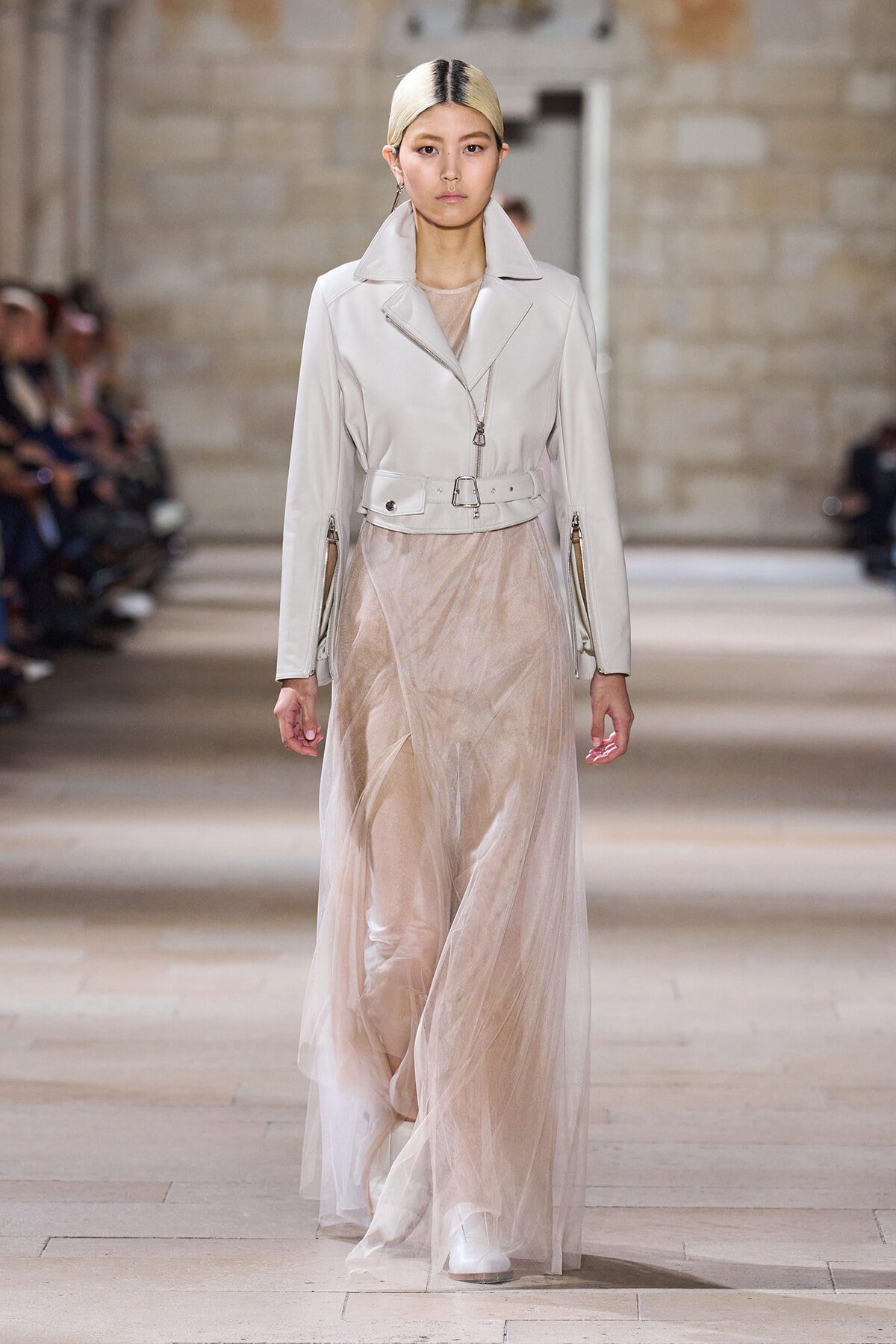 Model walks the runway in a pale leather jacket with a belt over a sheer beige gown.