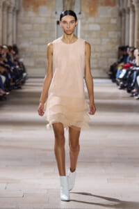Female model walks a runway in a pale peach sleeveless mesh dress with a layered ruffle hem and white ankle boots.