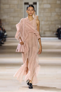 Model walking a runway in a pale pink, textured knit gown with a matching pink purse slung over the shoulder