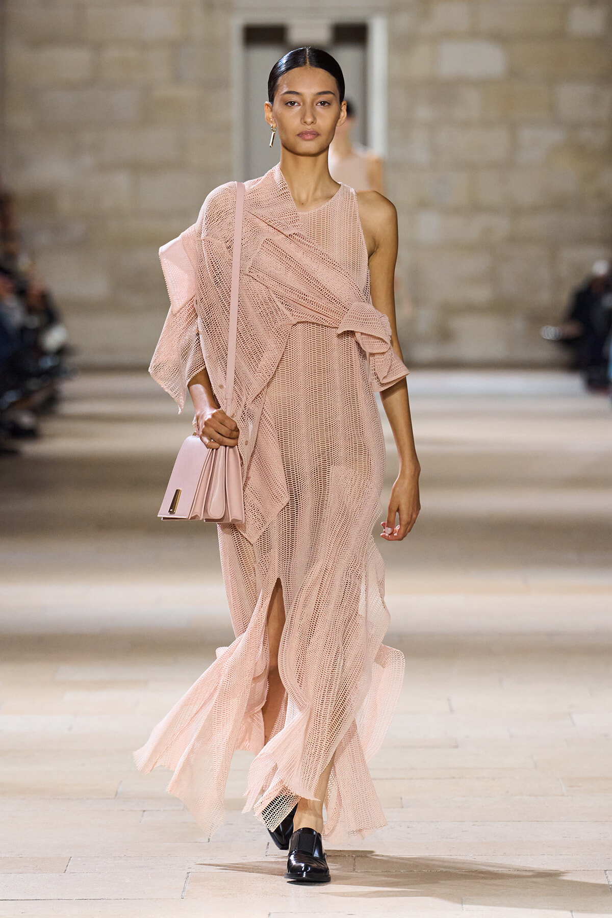 Model walking a runway in a pale pink, textured knit gown with a matching pink purse slung over the shoulder