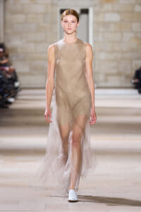 Model walking a runway in a sleeveless beige dress with a sheer tulle overlay and white flat shoes, in a fashion show setting.