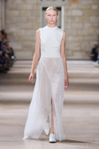 Model walking the runway in a sleeveless white high-neck dress with a sheer, flowing overlay.