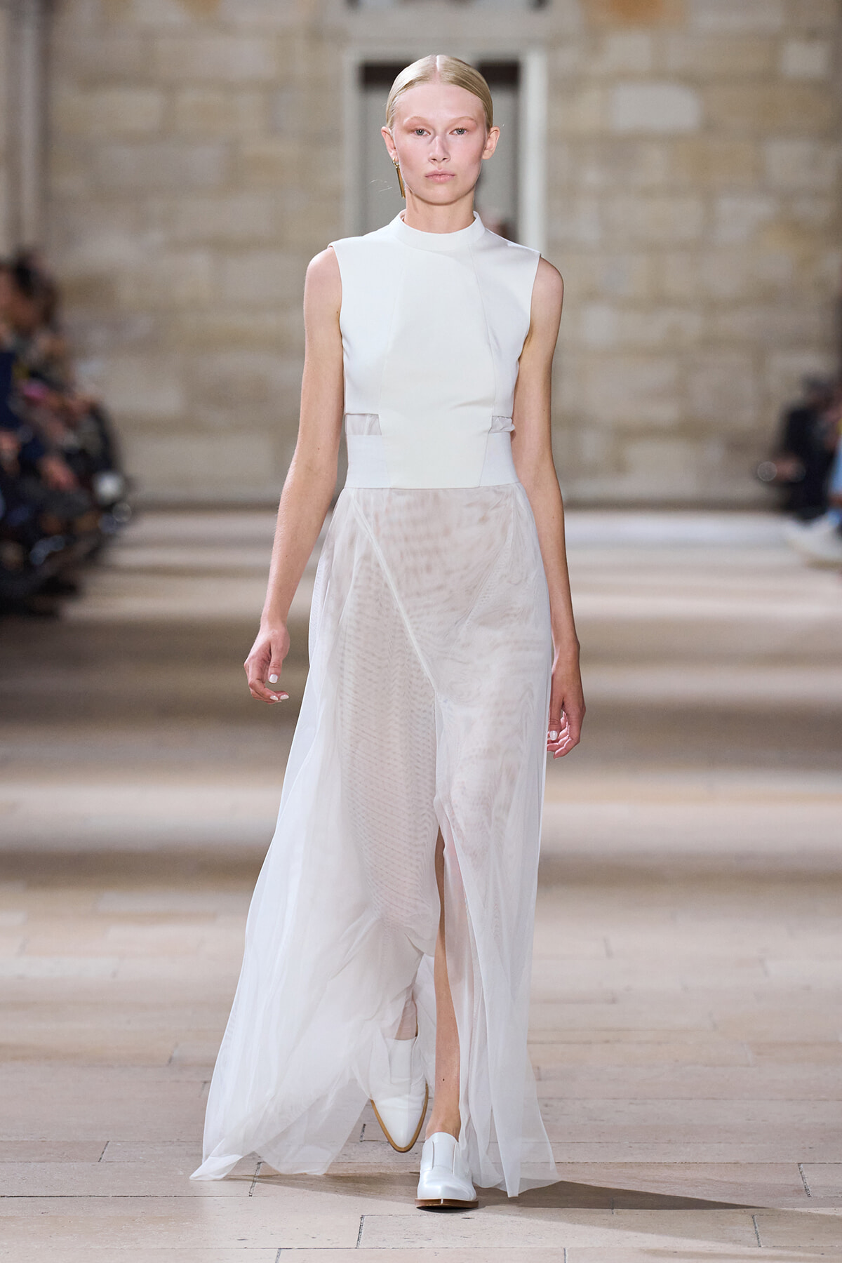 Model walking the runway in a sleeveless white high-neck dress with a sheer, flowing overlay.