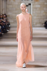 Model walking a sleeveless peach maxi dress down a fashion show runway in a stone-walled venue at the background audience scene.