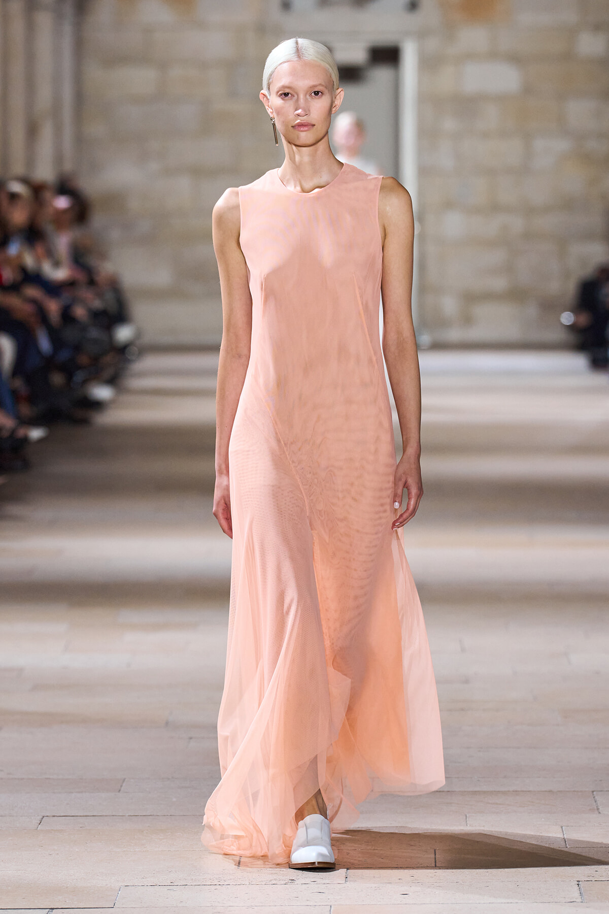 Model walking a sleeveless peach maxi dress down a fashion show runway in a stone-walled venue at the background audience scene.