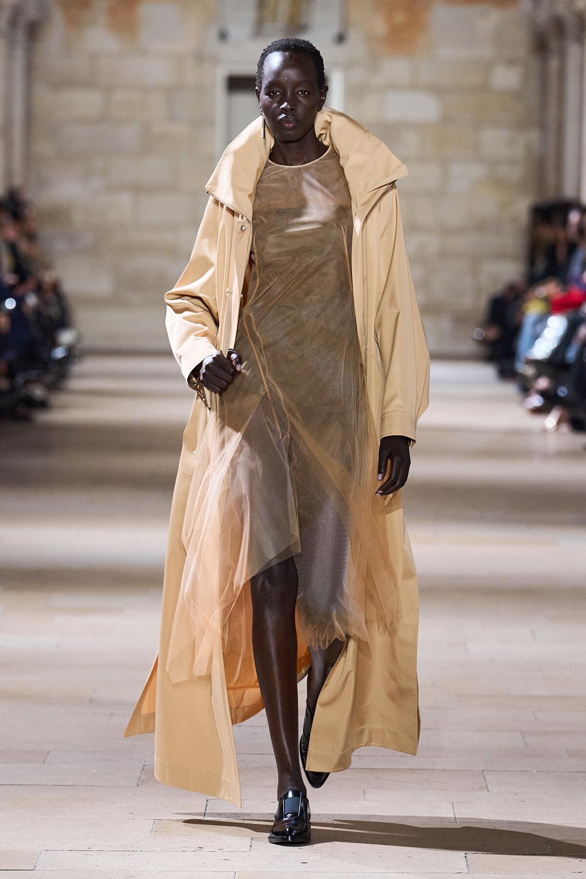 Model walks down runway in a beige trench over a sheer, bronze dress; black patent shoes and bold earrings visible in a fashion show setting.