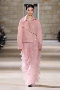 Model walking the runway in a pastel pink textured jacket over a sheer top and layered pink ruffle skirt at a fashion show