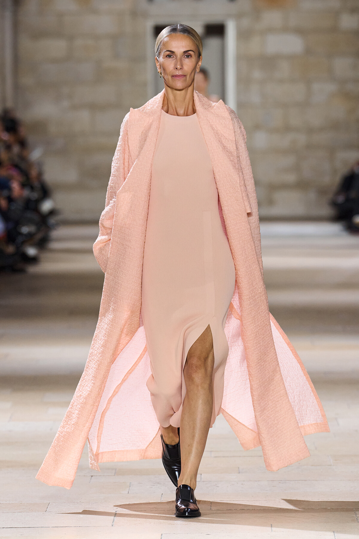 Runway model wearing a pale pink, sleeveless midi dress with a long textured pink cape, walking down the catwalk.