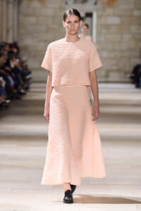 Model walking a fashion runway in a pale pink textured two-piece outfit: a short-sleeve cropped top and a long A-line skirt, set in a neutral venue.