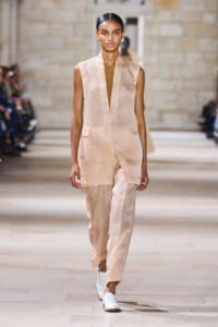 Model walks a fashion runway in a sleeveless beige blazer and matching wide-leg pants with white slip-on shoes.