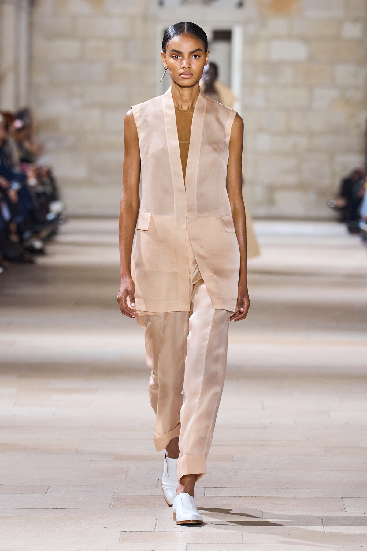 Model walks a fashion runway in a sleeveless beige blazer and matching wide-leg pants with white slip-on shoes.