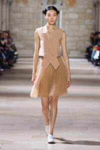 Model walking the runway in a beige sleeveless blazer with layered front and a knee-length textured tulle skirt.