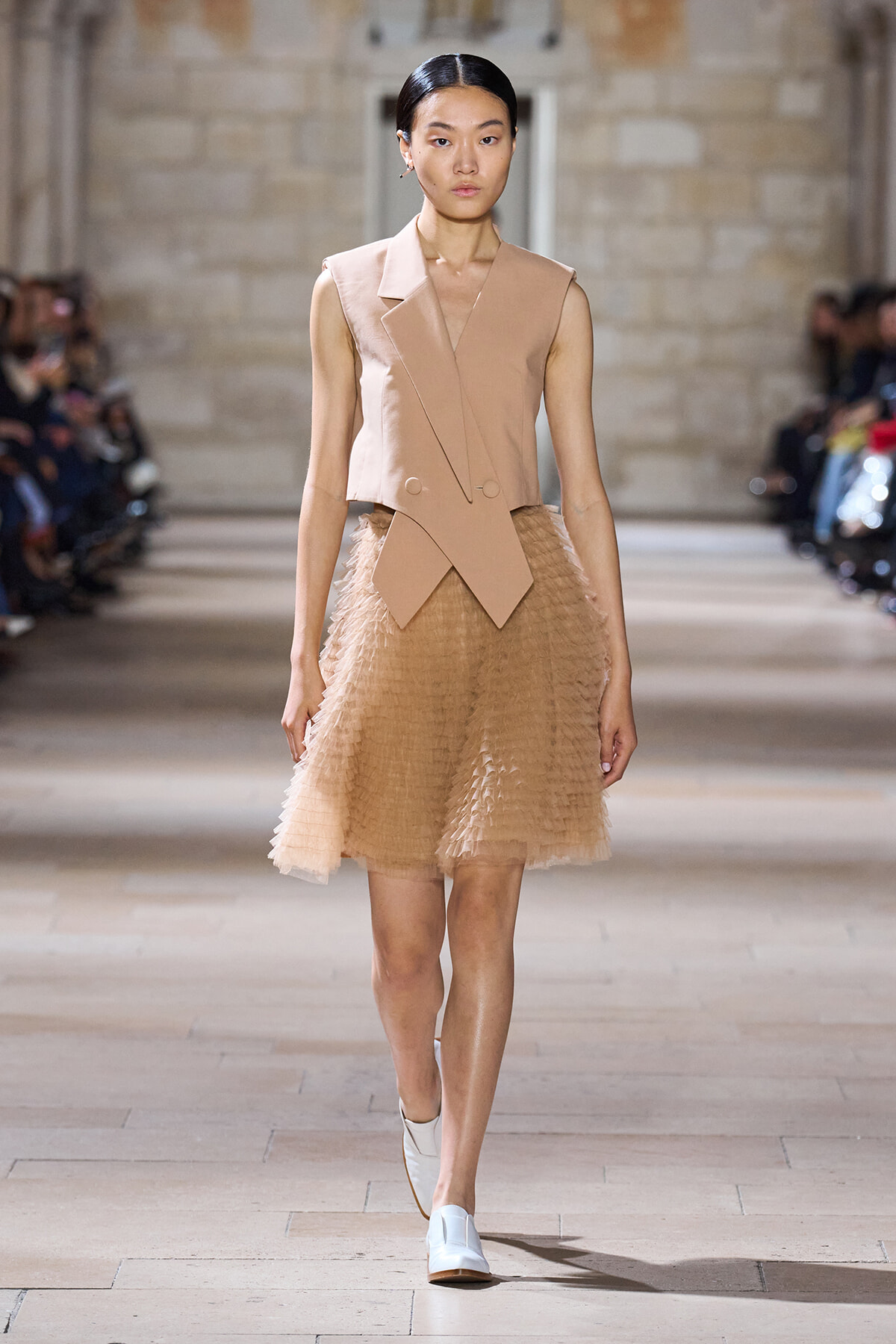 Model walking the runway in a beige sleeveless blazer with layered front and a knee-length textured tulle skirt.