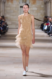 Model walks the runway wearing a knee-length beige sleeveless dress with layered ruffles and white loafers.