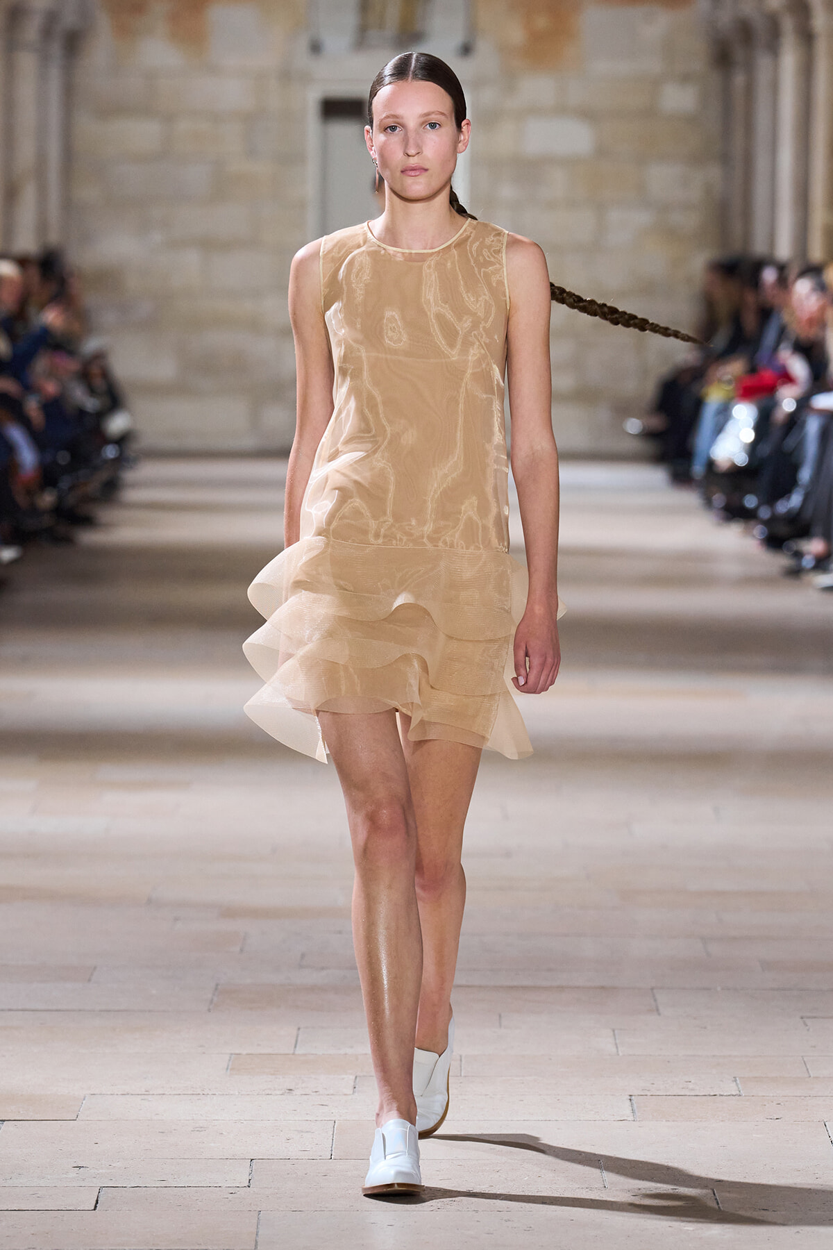 Model walks the runway wearing a knee-length beige sleeveless dress with layered ruffles and white loafers.