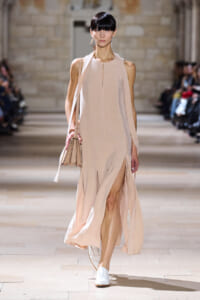 Model walks the runway in a pale pink sleeveless halter dress with a side slit and a beige handbag.
