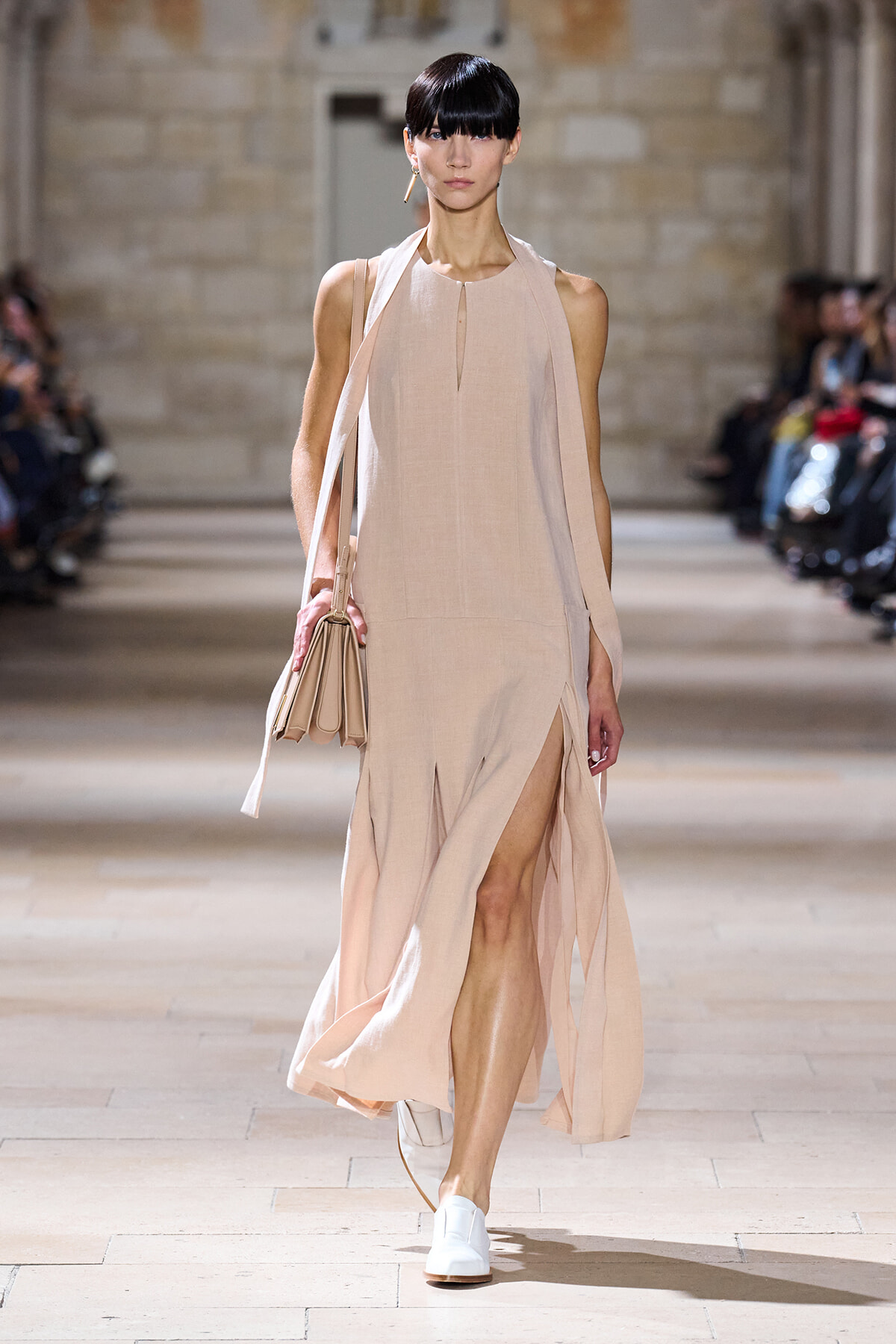 Model walks the runway in a pale pink sleeveless halter dress with a side slit and a beige handbag.