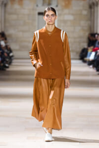 Runway model in a tan-brown leather-like jacket with white shoulder stripes and matching wide-leg pants, walking on a fashion show runway in a stone venue, wearing white shoes.