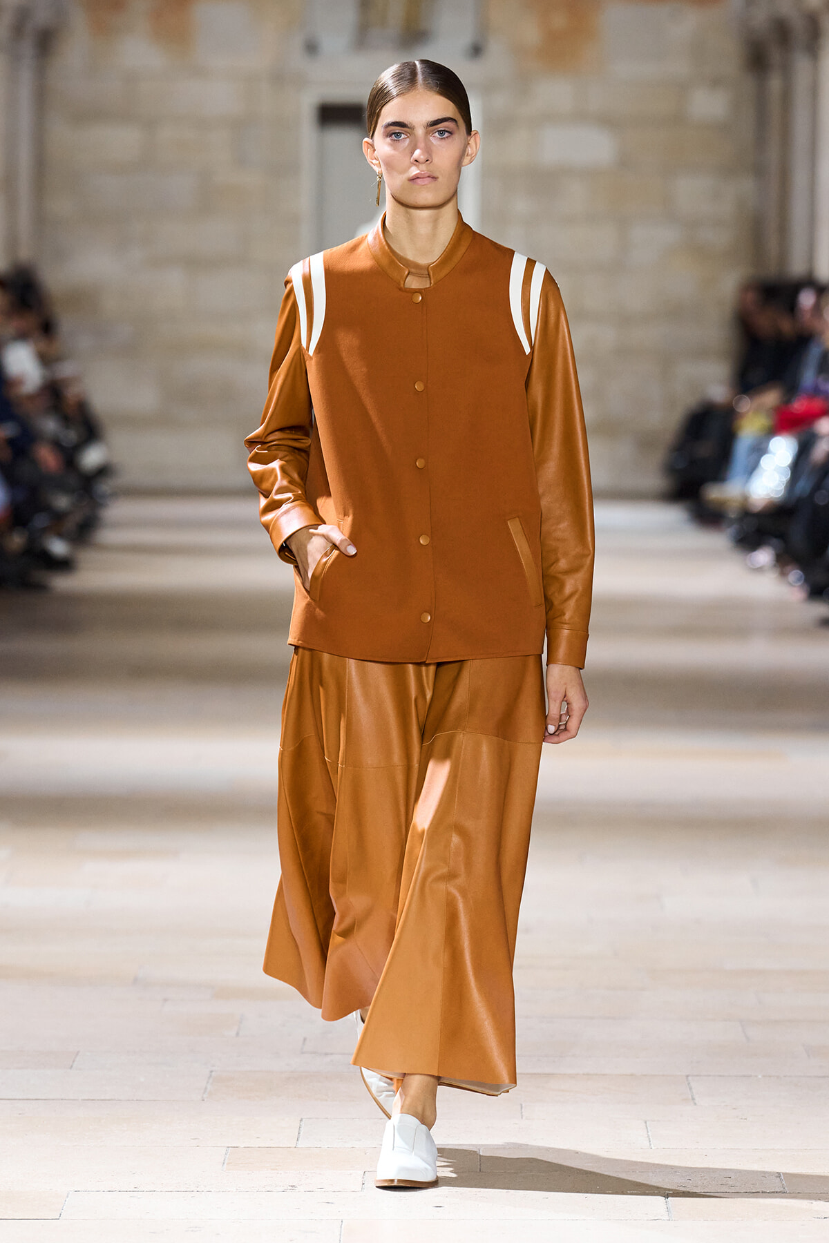 Runway model in a tan-brown leather-like jacket with white shoulder stripes and matching wide-leg pants, walking on a fashion show runway in a stone venue, wearing white shoes.