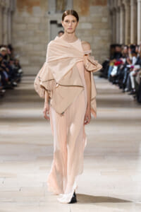 Model walking runway in a pale pink, off-shoulder, draped gown with oversized wrap detail and layered fabric folds.