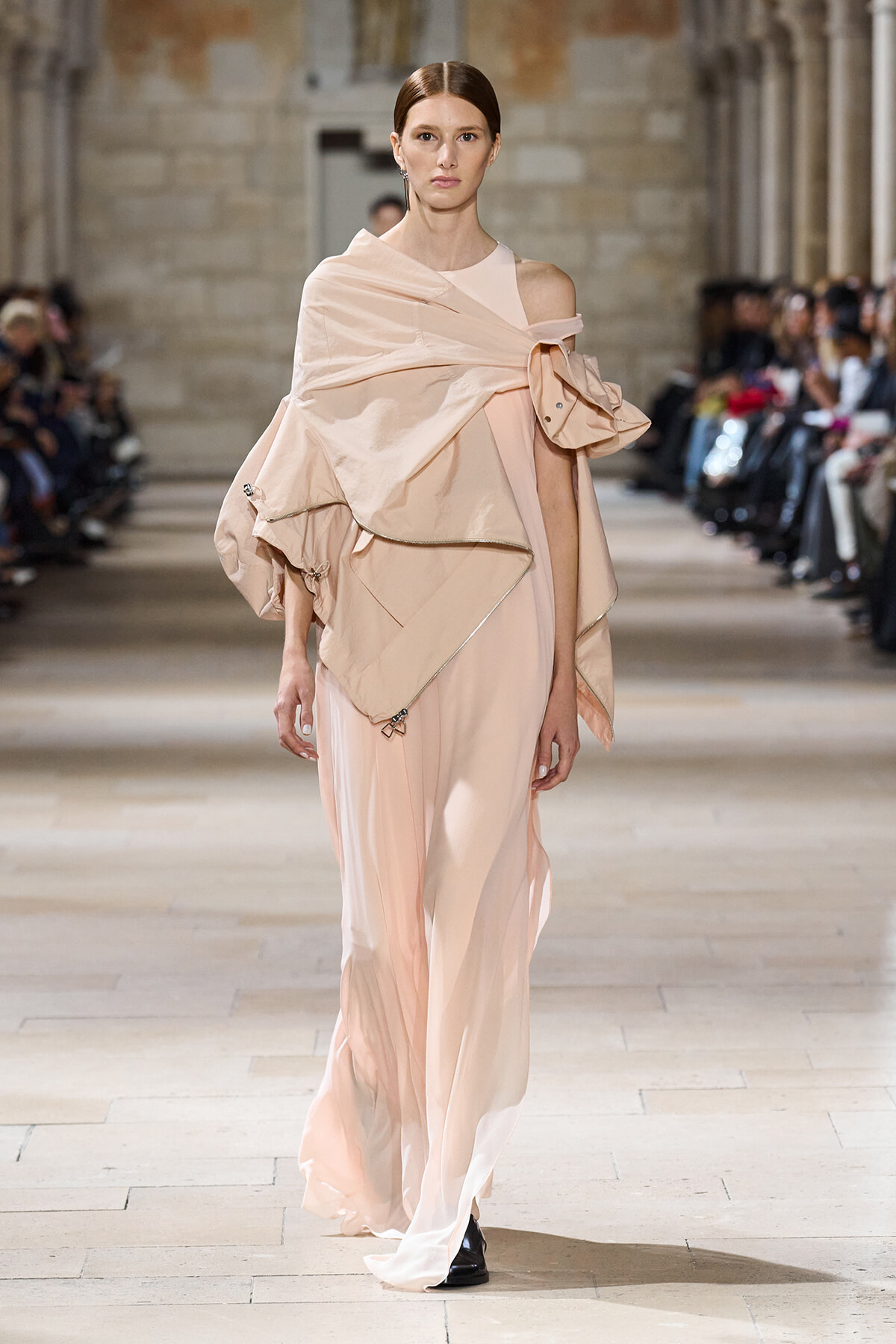 Model walking runway in a pale pink, off-shoulder, draped gown with oversized wrap detail and layered fabric folds.