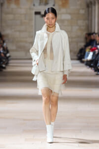 Model on a runway in an all-white ensemble: oversized jacket, sheer top, layered mini skirt, and white ankle boots with a white bag on her shoulder