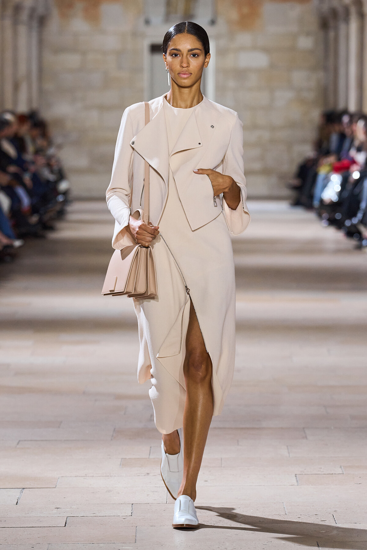Model walking a cream trench dress with draped lapels, carrying a structured pale purse on a fashion runway.